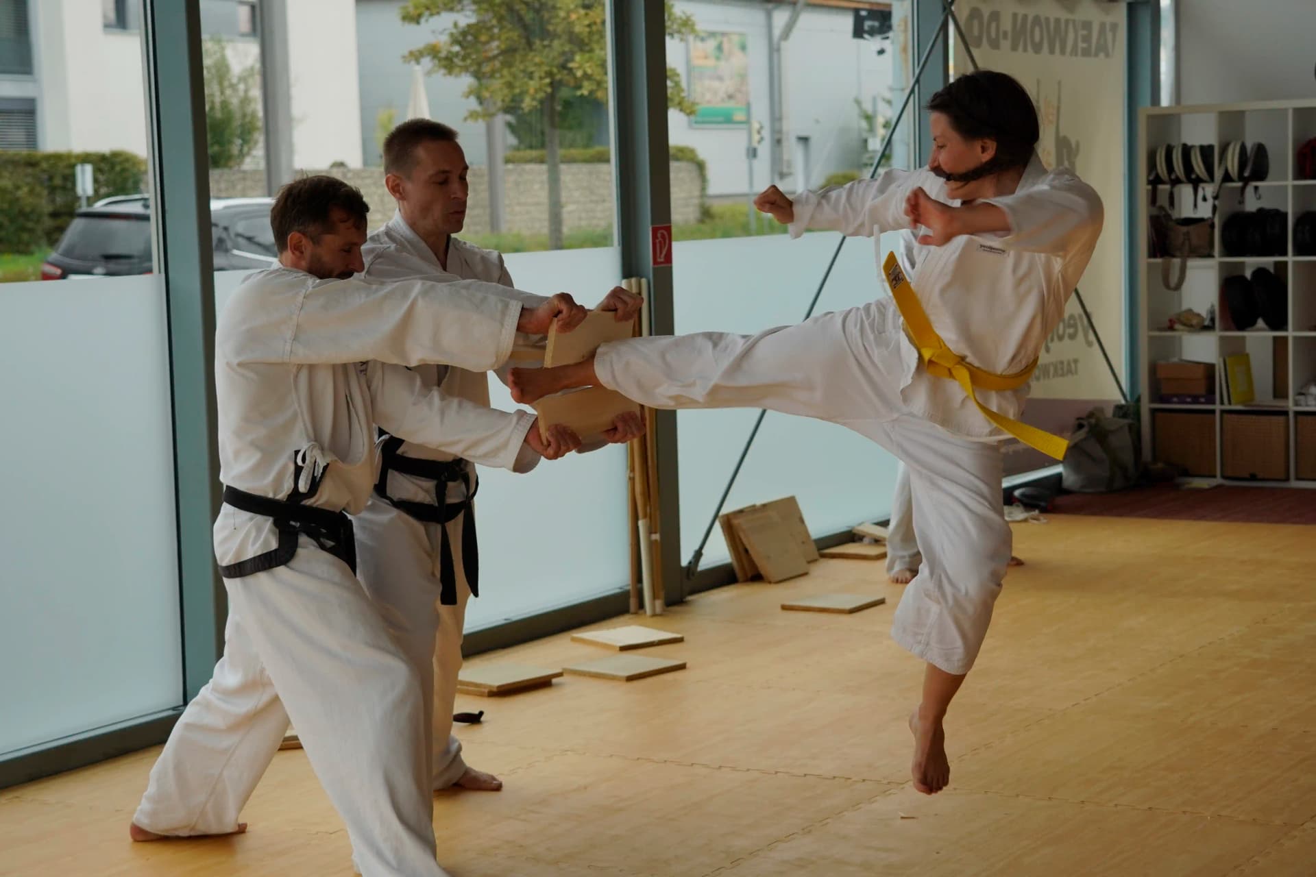 Taekwondo-Schüler führt während der Gürtelprüfung einen Yop Chagi (Seitwärtskick) als Bruchtest aus. Zwei Schulleiter halten das Brett stabil. Fokus auf Konzentration und Technik beim Bruchtest.