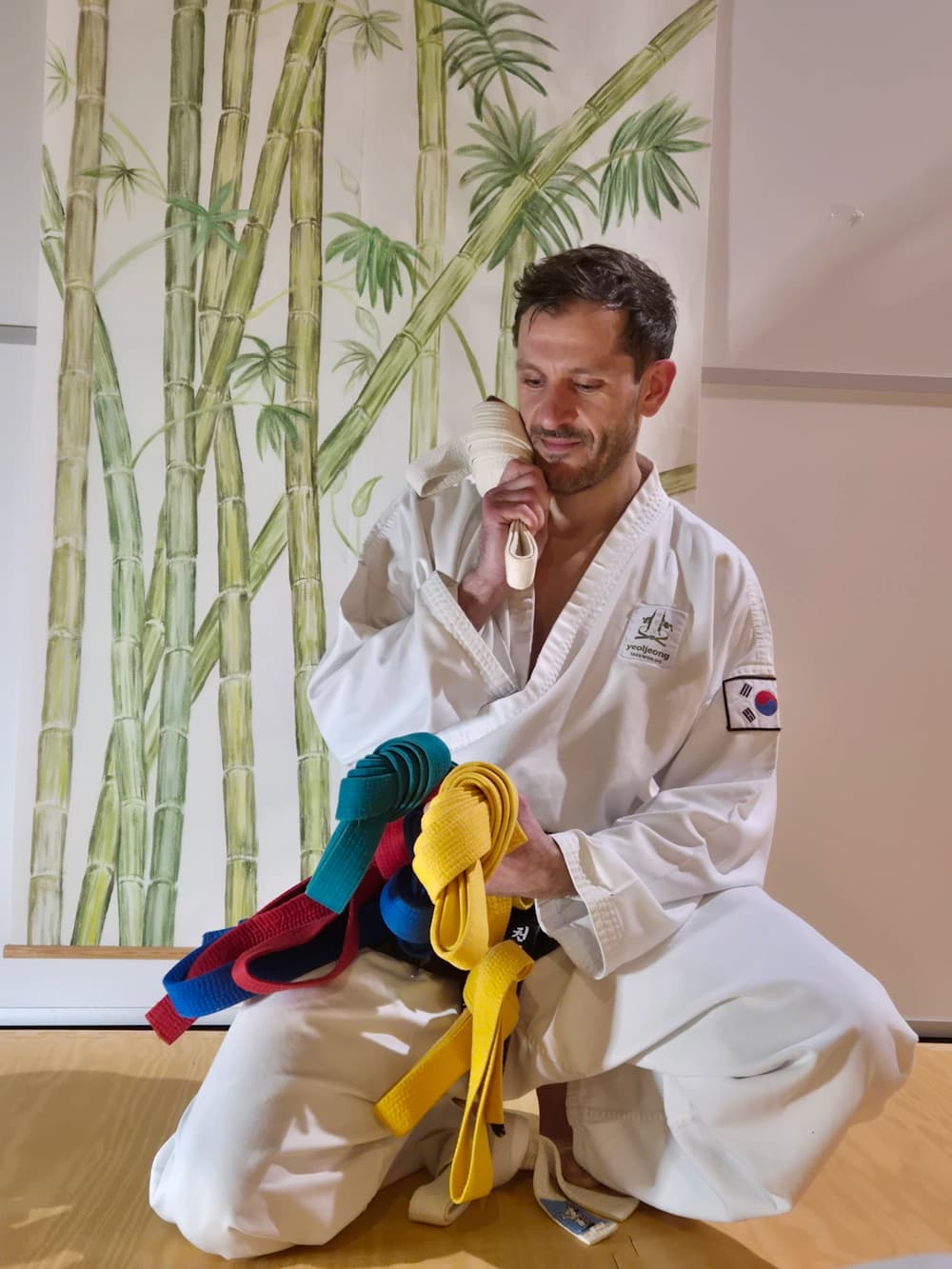 Authentisches Foto eines Kampfsport-Schulleiters, der mit ratlosem, fragendem Blick verschiedene farbige Gürtel in den Händen hält. Symbolisiert die Unsicherheit bei der manuellen Trainingsplanung und Prüfungszulassung ohne Software-Unterstützung.